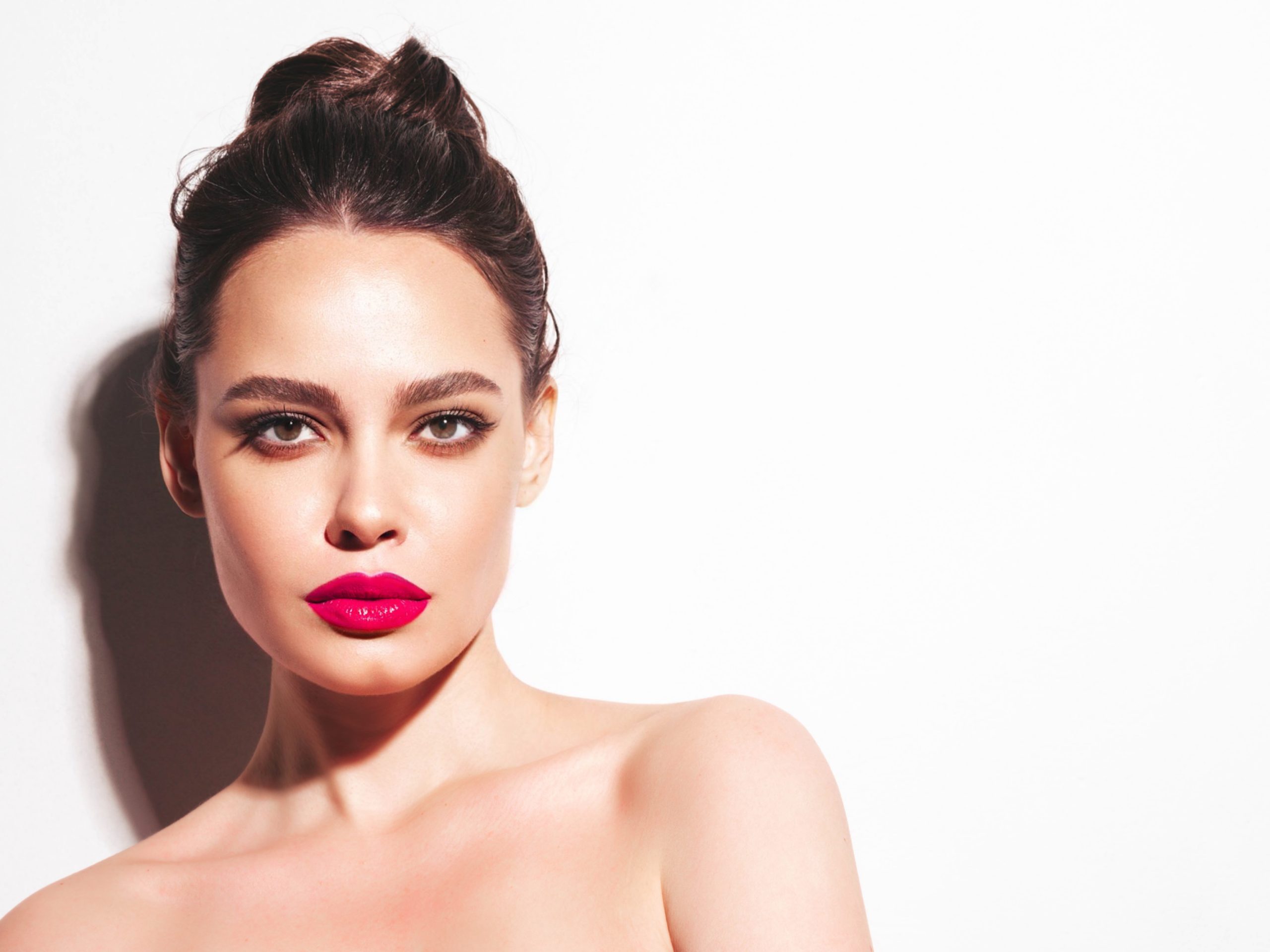 Fashion beauty portrait of young brunette woman with evening stylish makeup and perfect clean skin. Sexy model with hair in a bun posing in studio near white wall. With pink bright natural lips